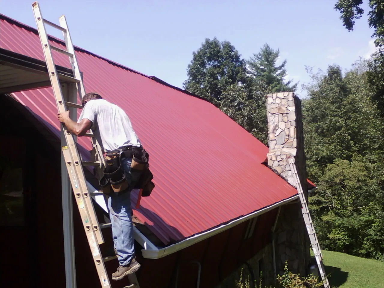 Roofer on a ladder beside a red metal roof and stone chimney — Soffit & Fascia Repair by Roofing Brothers in Burke